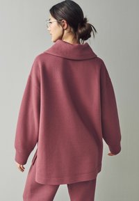 Üks suuruses pullover, mis on valmistatud tekstuursest sügava roosa kangast, millel on lai kaelus. Omab langenud õlgu ja külgmisi lõikeid liikumisvabaduse saavutamiseks.