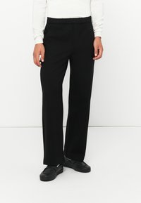 Schwarze, weit geschnittene Hosen mit einem elastischen Bund und seitlichen Taschen, aus glattem Stoff. Getragen mit schwarzen Slippern.