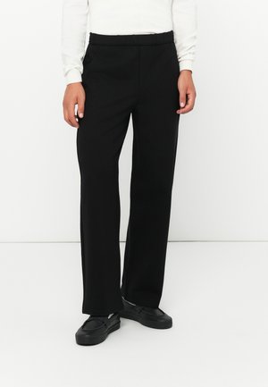 Schwarze, weit geschnittene Hosen mit einem elastischen Bund und seitlichen Taschen, aus glattem Stoff. Getragen mit schwarzen Slippern.