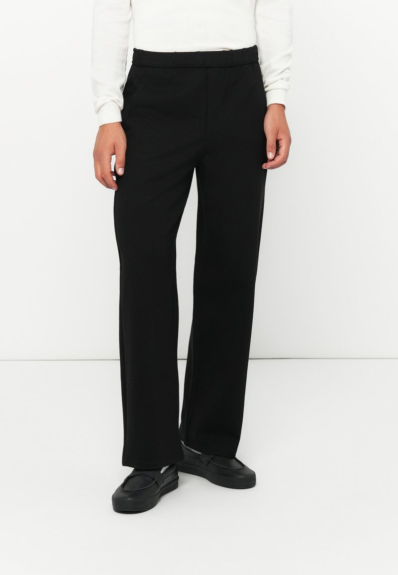 Schwarze, weit geschnittene Hosen mit einem elastischen Bund und seitlichen Taschen, aus glattem Stoff. Getragen mit schwarzen Slippern.