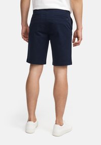 Shorts en coton marine avec coupe droite, deux poches arrière et un ourlet net. Portés avec des baskets blanches, montrant un style décontracté et une coupe relax.