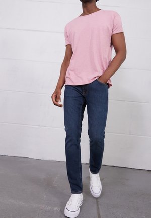 Mężczyzna idący, ubrany w jasnoróżowy t-shirt z krótkim rękawem, ciemnoniebieskie dżinsy i białe trampki, z prawą ręką w kieszeni, na tle białej ściany.