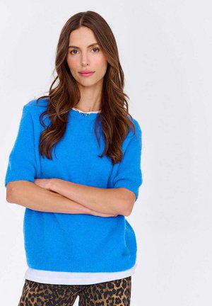 Modrá krátkopesťová svetrová s jemnou textúrou, preložená cez bielu košeľu, doplnená nohavicami s leopardím vzorom. Neformálny a uvoľnený dizajn.