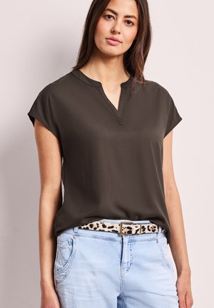 Kvinde iført mørkeblå olivengrøn kortærmet top stoppet ned i lyseblå jeans med leopardprint bælte, stående mod en ensfarvet baggrund.