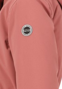 Giacca leggera rosa con spalline che presenta una patch circolare in tessuto con il logo "ZIGZAG" e il testo. Texture liscia, design pulito e aspetto sportivo.