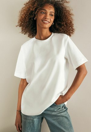 Witte t-shirt met korte mouwen en ronde hals, in een relaxte pasvorm, gemaakt van een gladde stof. Heeft een subtiele gebogen zoom en wordt gedragen met lichtblauwe spijkerbroek.