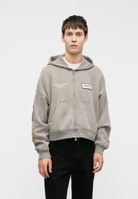 Hallhalli halli, millel on lühendatud disain, tikitud detailid, kaks eesmist taskut ja soonikkoes varrukad, kantud mustade pükstega.