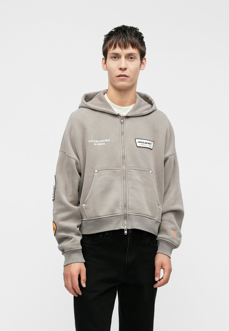 Hallhalli halli, millel on lühendatud disain, tikitud detailid, kaks eesmist taskut ja soonikkoes varrukad, kantud mustade pükstega.