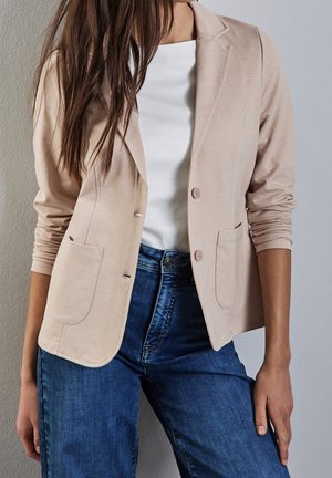 Kvinde iført beige blazer over hvid skjorte og blå jeans står mod en lysegrå væg med en arm afslappet ned langs siden.