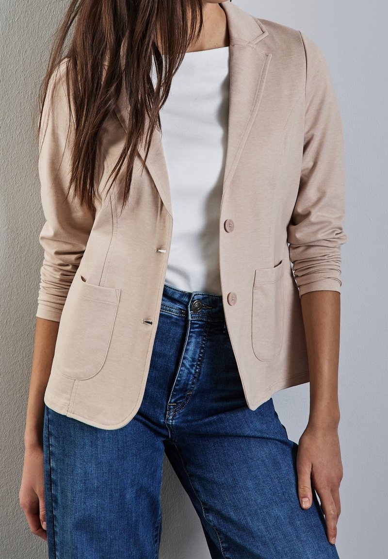 Femme portant un blazer beige sur une chemise blanche et un jean bleu, debout contre un mur gris clair avec un bras détendu le long du corps.