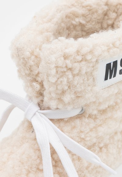 Botim de tornozelo bege, fofo, com uma superfície texturizada, fecho de atacadores brancos e uma etiqueta de marca com letras pretas.