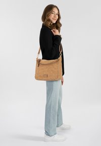 Braune Lederschultertasche mit strukturiertem Finish, angereichert mit einem verdrehten Tragegurt und einem Logo-Schild. Tragen zu hellblauen Jeans und weißen Sneakers.