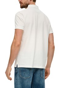 Weißes Polohemd mit kurzen Ärmeln und Kragen, mit geradem Saum und seitlichen Schlitzen; kombiniert mit blauen Jeans.