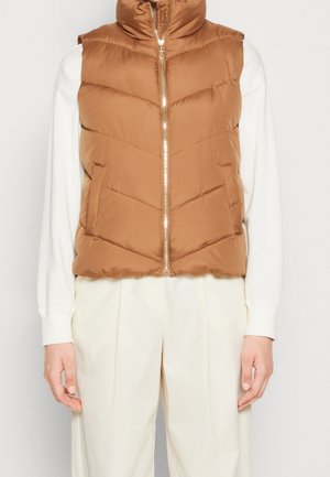 Veste sans manches matelassée marron à fermeture éclair portée sur un haut blanc à manches longues et un pantalon crème taille haute.