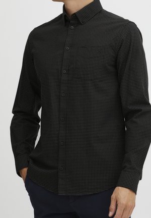 Mand iført en tætsiddende sort skjorte med små tern og lange ærmer, hænder delvist i lommerne på marineblå bukser.