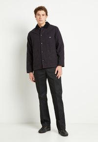 Giacca in cotone blu navy con chiusura a bottoni, quattro tasche frontali e colletto in corduroy, abbinata a pantaloni neri e scarpe scure.