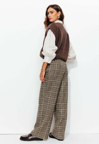 Gilet en polaire marron porté sur une blouse à carreaux crème avec des manches bouffantes, associé à un pantalon large à carreaux marron et beige, chaussures noires.