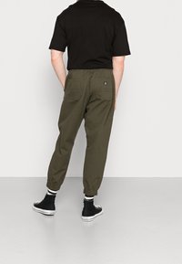 Persoon in olijfgroene joggerbroek met achterzakken, zwart T-shirt, gestreepte sokken en zwarte hoge Converse sneakers, staand op een grijze vloer.