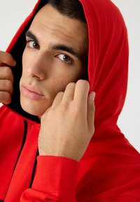 Sudadera de algodón roja con capucha ajustable, llevada por una persona, que presenta una textura suave y puños acanalados. El fondo es neutro.