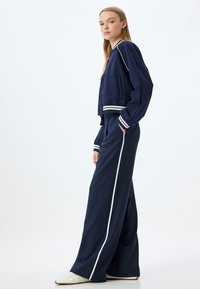 Navy Sportjacke mit weißen Streifen, kombiniert mit passenden weit geschnittenen Hosen. Der Stoff wirkt glatt, mit einem lockeren Schnitt und einem elastischen Bund.