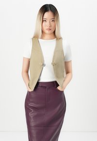 Beige vest med en figursydd design, med to frontspenner. Kombinert med et lilla skinnskjørt, glatt tekstur og rett silhuett.