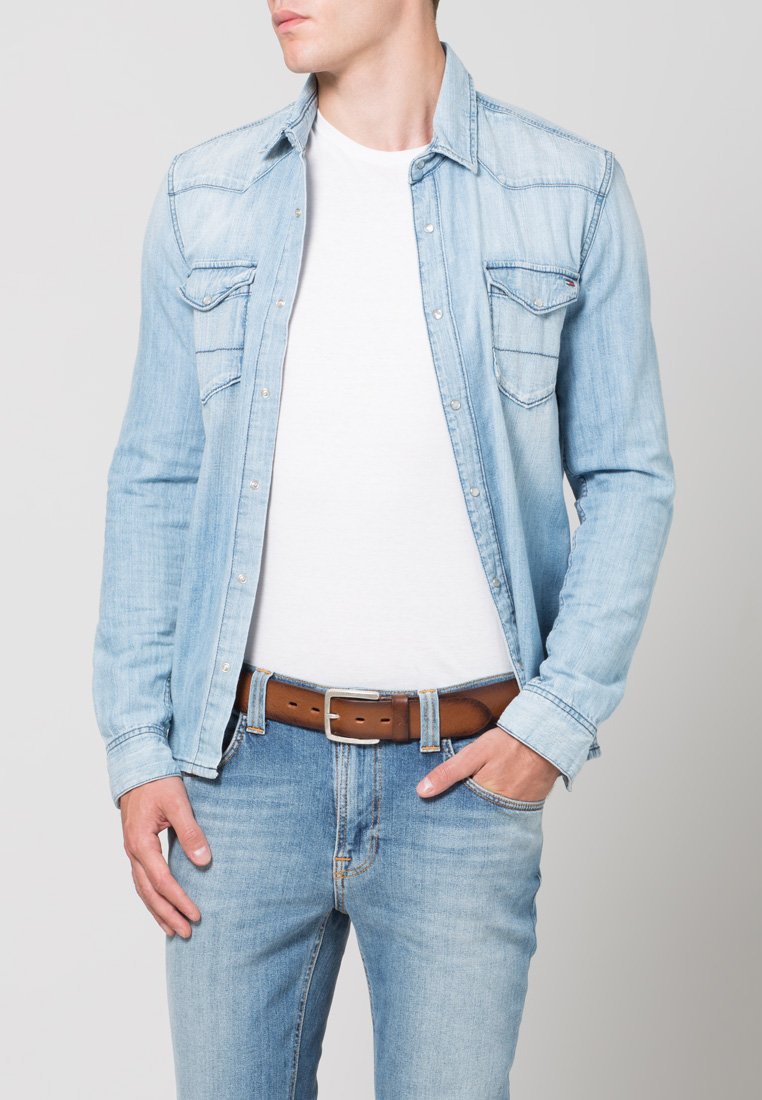 Camisa de denim azul claro con dos bolsillos en el pecho y cierre de botones. Usada sobre una camiseta blanca, combinada con jeans azul claro y un cinturón marrón.