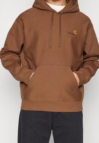 Felpa con cappuccio marrone realizzata in tessuto morbido e testurizzato, dotata di una tasca frontale e di un piccolo logo ricamato sul petto.