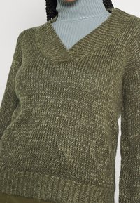 Maglione lavorato a maglia verde oliva con scollatura a V, tessuto testurizzato e polsini a coste. Presenta un taglio ampio e un motivo a maglia grossa.