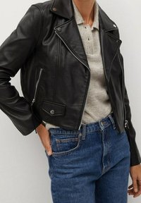 Zwarte leren motorjack met asymmetrische rits, zilverkleurige accenten en zijzakken, gecombineerd met een lichtgrijze trui en blauwe jeans.