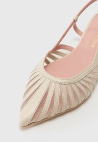 Béžová slingback obuv s špicatou špičkou, so sieťovými panelmi a vertikálnymi pruhmi. Mäkká ružová stielka s zlatým brandingom.