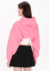 Sudadera corta rosa con capucha y mangas oversize, combinada con una mini falda plisada negra. Textura suave, diseño casual.