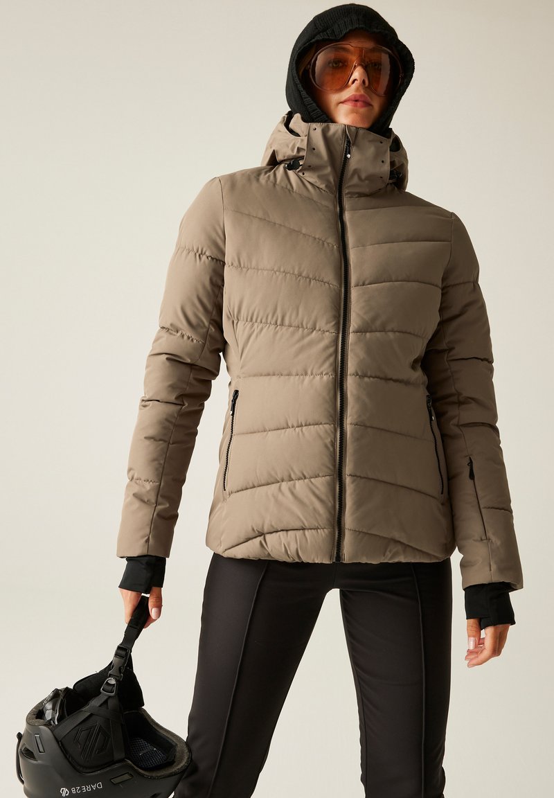 Veste matelassée beige avec des coutures verticales, des poches zippées et une capuche amovible ; associée à un pantalon noir et tenant un casque noir.
