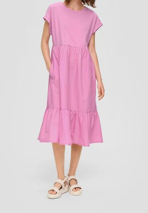 Kobieta ubrana w luźną, różową sukienkę midi z krótkim rękawem, falbaną na dole, bocznymi kieszeniami i sandałami na białych plecionych paskach.