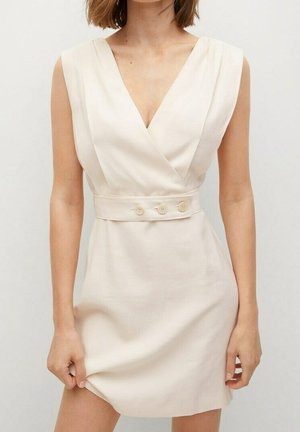 Robe portefeuille beige sans manches avec décolleté en V et large ceinture ornée de trois boutons, portée par une femme tenant l'ourlet de la robe.