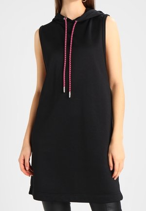 Robe noire sans manches à capuche avec cordons roses et blancs, portée sur un pantalon noir, visible du cou au milieu de la cuisse.