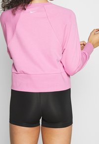 Femme portant un haut à manches longues rose de Nike et un short de sport noir, vue de dos sur un fond uni.