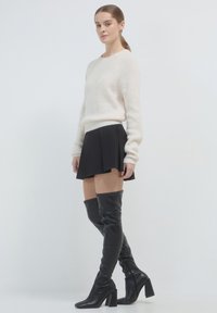 Cremfarbener Strickpullover, plissierter schwarzer Minirock und schwarze überkniehohe Lederstiefel mit klobigen Absätzen, vor einem neutralen Hintergrund.