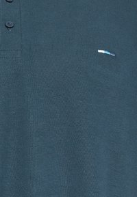 Polo bleu sarcelle foncé en tissu texturé, avec deux boutons noirs près du col et un petit logo rayé horizontal sur la poitrine.