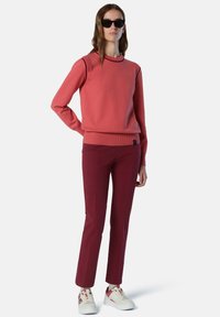 Jersey rojo con cuello y puños acanalados, combinado con pantalones burdeos. La modelo lleva gafas de sol negras y zapatillas blancas con toques rojos.