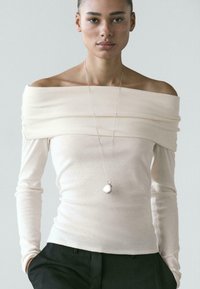 Kremowy, gładki top z długim rękawem i dekoltem odsłaniającym ramiona, z warstwowym wzorem tkaniny. Do tego srebrny naszyjnik z zawieszką.