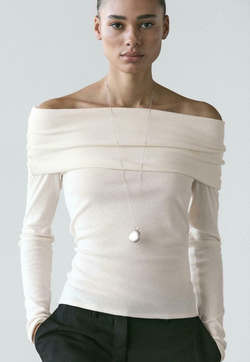 Kremowy, gładki top z długim rękawem i dekoltem odsłaniającym ramiona, z warstwowym wzorem tkaniny. Do tego srebrny naszyjnik z zawieszką.