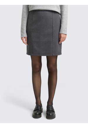 Femme portant une mini-jupe gris pied-de-poule, des collants noirs transparents, des mocassins noirs et un pull gris à manches longues, debout devant un fond uni.