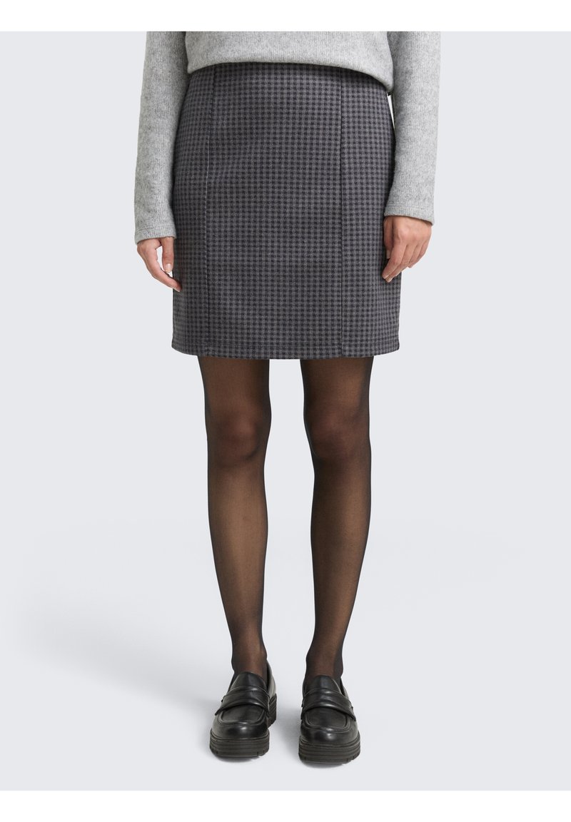 Femme portant une mini-jupe gris pied-de-poule, des collants noirs transparents, des mocassins noirs et un pull gris à manches longues, debout devant un fond uni.