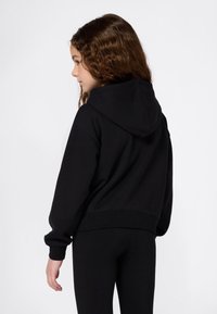 Niña joven con cabello castaño ondulado, usando una sudadera con capucha negra y pantalones negros, mirando hacia la izquierda contra un fondo blanco.