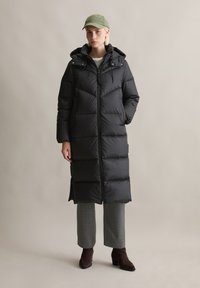Zwarte puffy parka met capuchon, voorzien van een ritssluiting aan de voorkant, diagonale gestikte patroon en zijzakken. Model draagt grijze geruite broek en bruine laarzen.