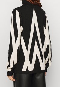 Schwarz-weißer gestrickter Pullover mit hohem Kragen, ausgestattet mit einem Zickzackmuster, langen Ärmeln und lockerer Passform. Strukturiertes Material.