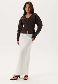 Brun stickad cardigan med V-ringning och dragkedja fram, ihop med en figurnära vit maxikjol. Modellen bär svarta remsandaler.