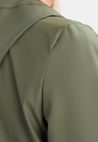 Olivengrøn hættejakke med en glat, blank overflade. Har strukturerede skuldersømme og en blød finish, der understreger et moderne design.