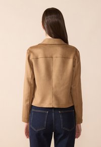 Veste en suédine beige avec un col, un design ajusté et des manches longues ; portée avec un jean bleu foncé, mettant en valeur les détails au dos et les coutures.
