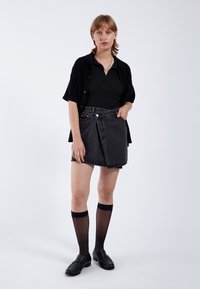 Zwarte denim wikkelrok met een sluiting van knopen aan de voorkant, gecombineerd met een zwart geribbeld topje en een oversized zwart overhemden. Kniehoge doorschijnende sokken en loafers.
